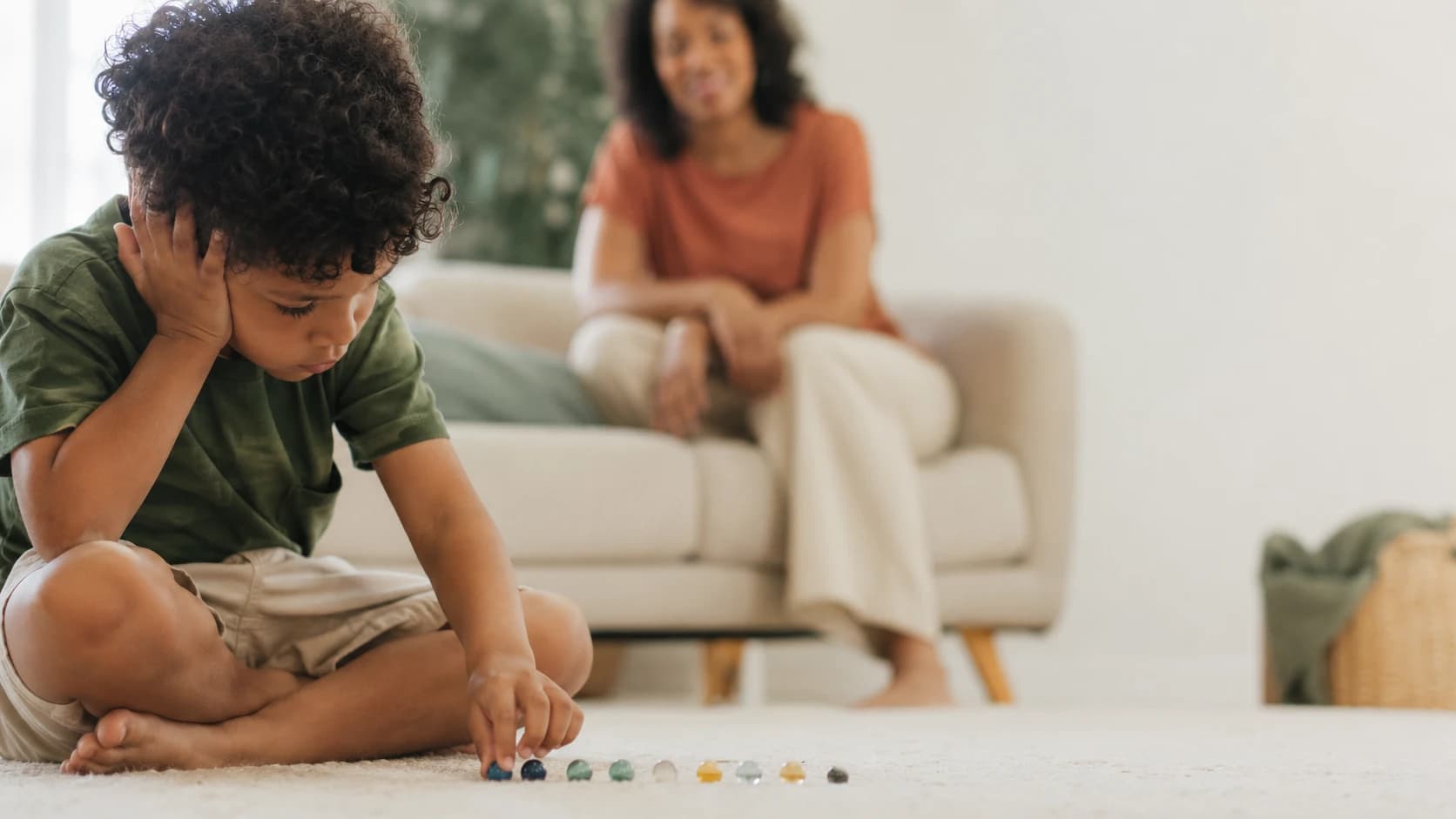 Criança concentrada organizando objetos em fileira, mãe observando com carinho ao fundo.